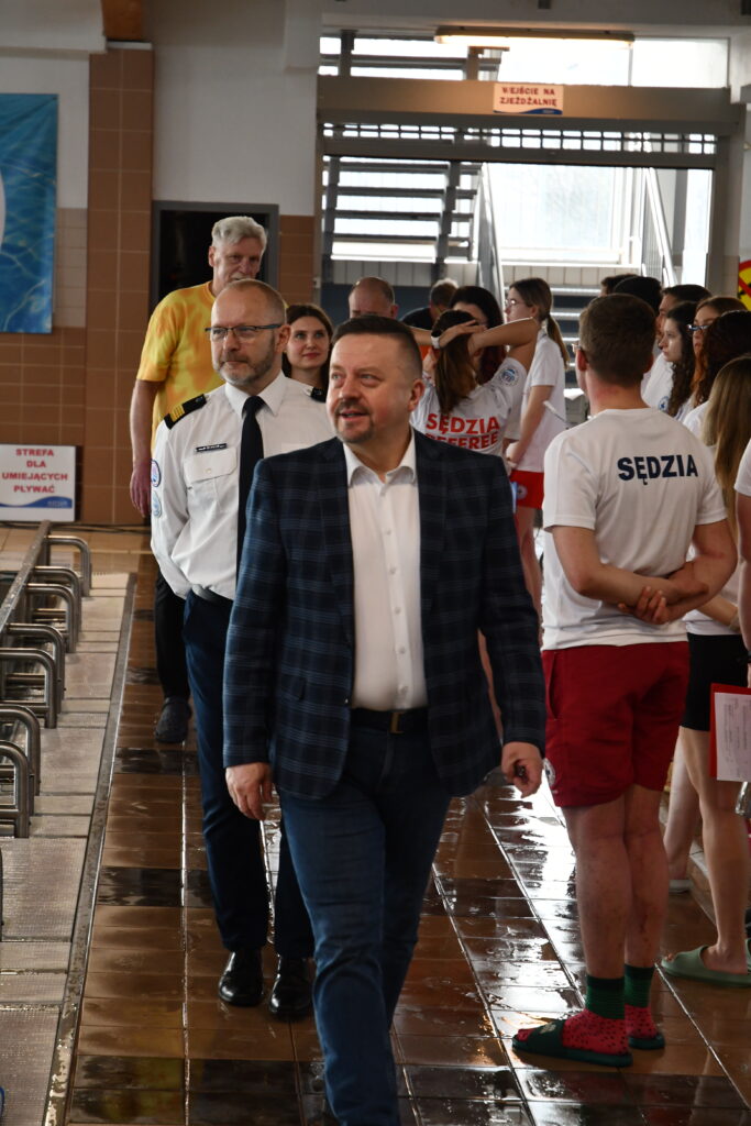 Mężczyźni idą obok basenu, jeden w marynarce, drugi w koszuli z pagonami, w tle sędziowie.